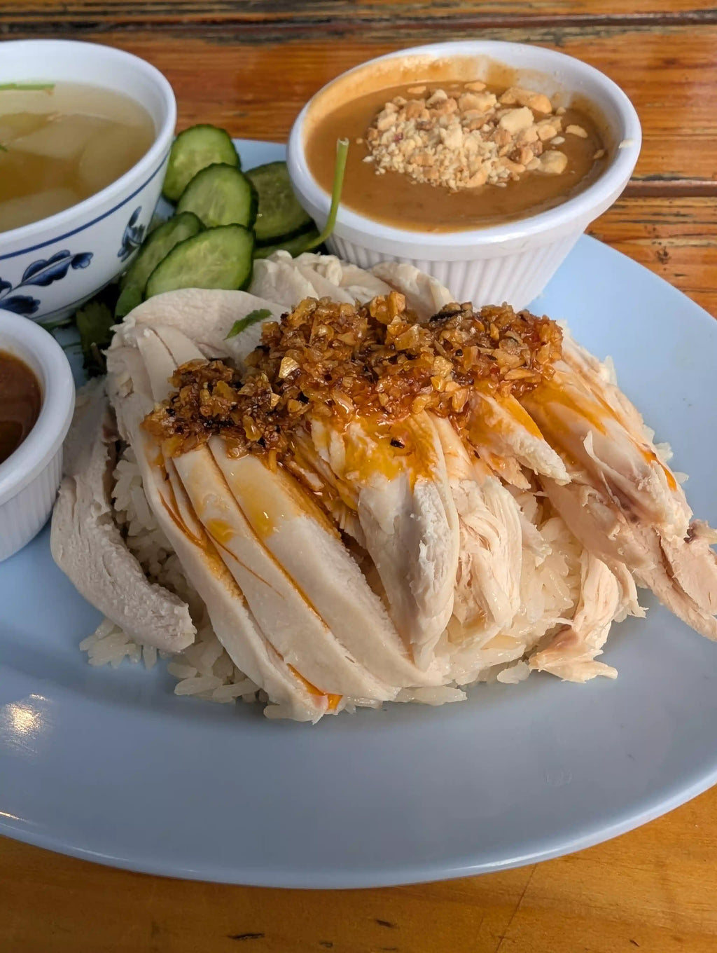 Plated dish of rice with chicken, chili oil, and a side of soup and pickles on a wooden table.