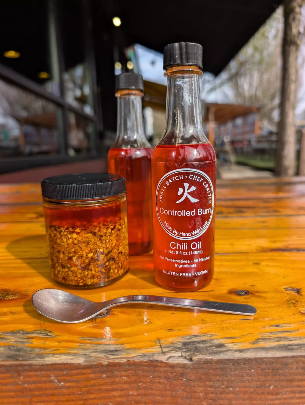 Two bottles of 'Controlled Burn' chili oil and a jar of chili flakes on a wooden table.